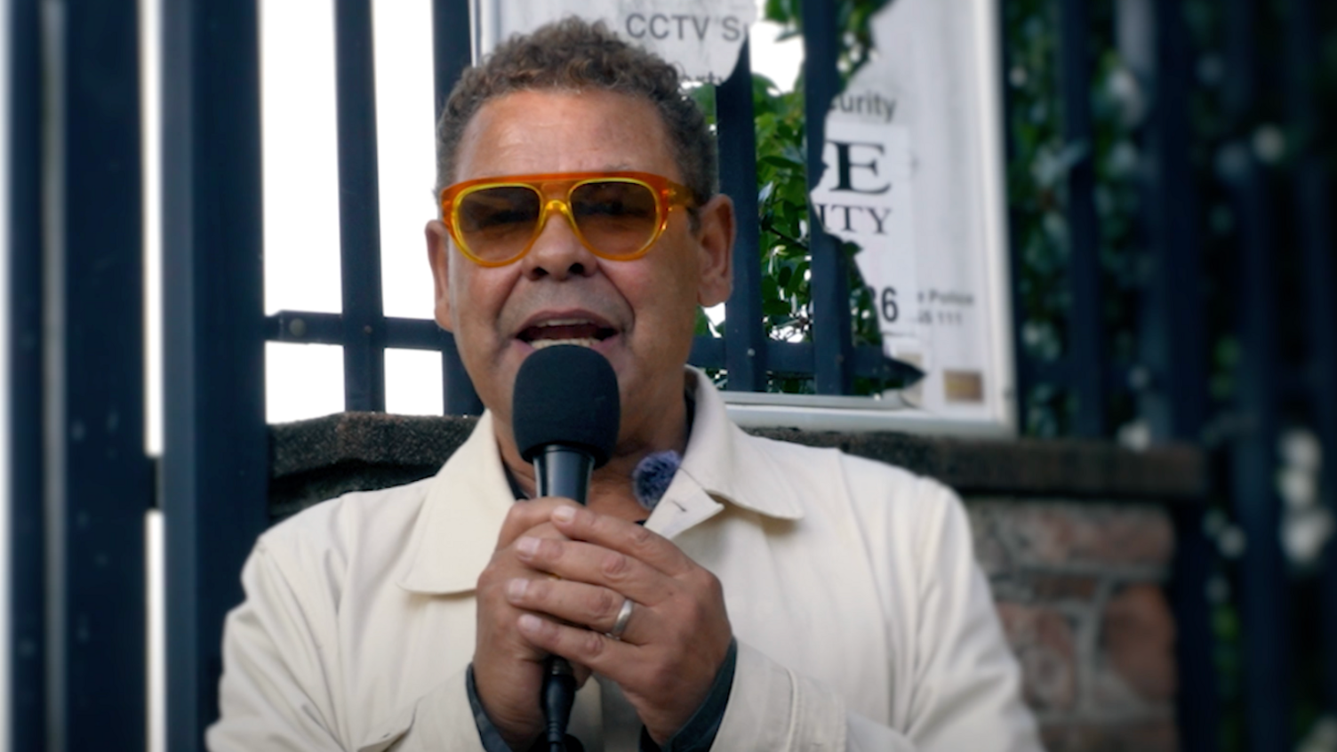 Craig Charles has short brown curly hair and is wearing brown-framed glasses and a cream coat. He is speaking into a microphone.