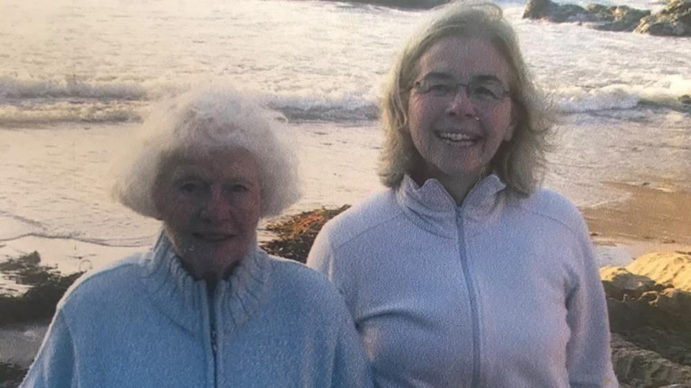 An old photo of Sheila and Bern who are both wearing light-coloured jumpers while standing on a beach with the sea in the background.