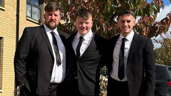 Three man in dark suits and black ties, standing outside a house, with parked cars behind them.