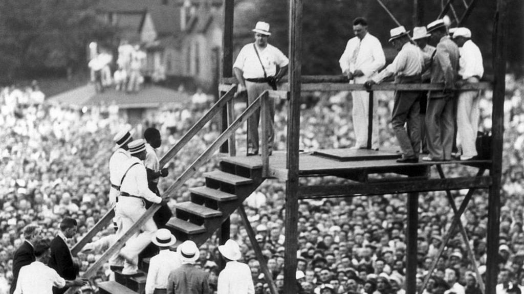 Men stand on wooden gallows, as another man is escorted up some steps to the stage from which he will be executed.