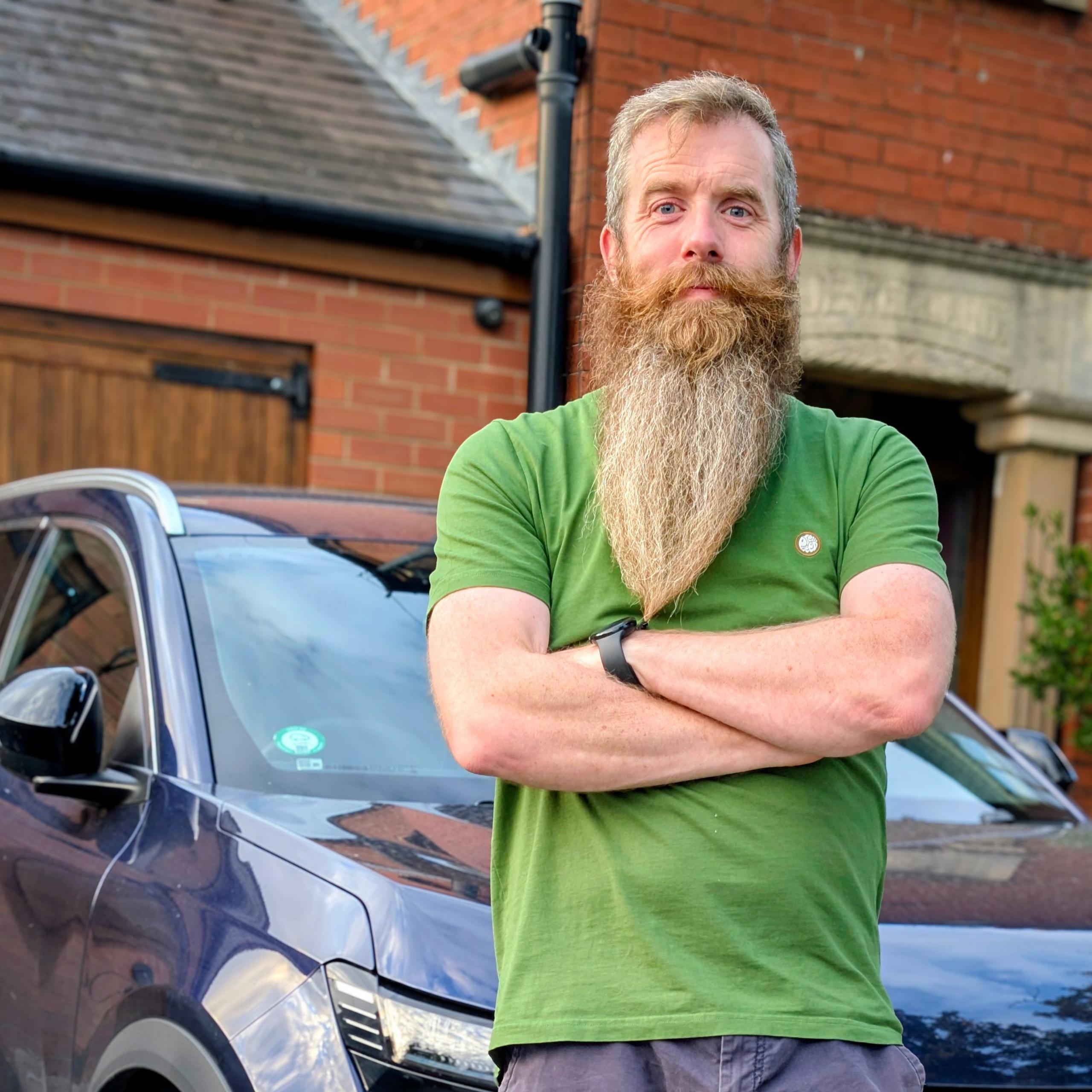 Mark Quigg, a man with a long beard in a green T-shirt and grey shorts, stands in front of a dark blue Renault, which is parked on a driveway in front of a house
