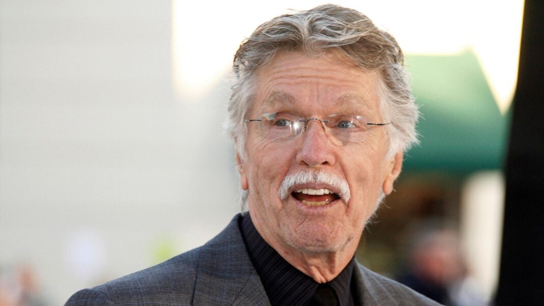 Actor Tom Skerritt is seen with his mouth open, apparently talking, and smiling. He is wearing a black buttoned-up shirt, rimless glasses and a grey suit coat. He appears to be outside.