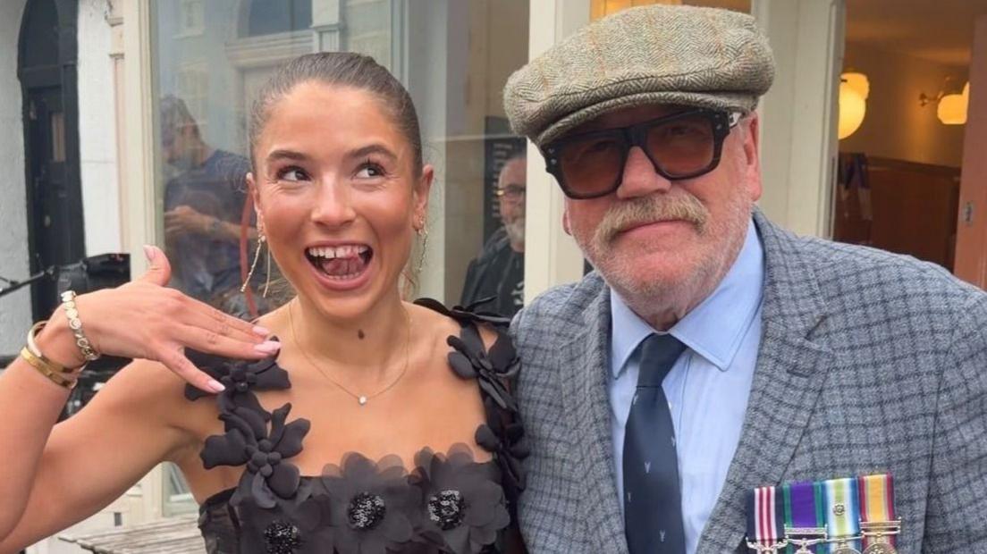 Ray Winstone has a big moustache, thick rimmed glasses and a blue blazer with military medals on the lapel, he is seen with Molly Ridge who is wearing a black dress made of fabric flowers and acts shocked to see the film star