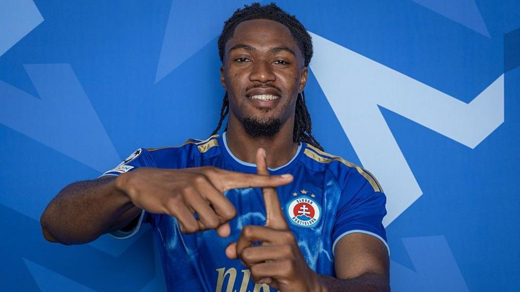 A man places one finger over the other to create a plus symbol while smiling. The move mimicks a goal celebration.