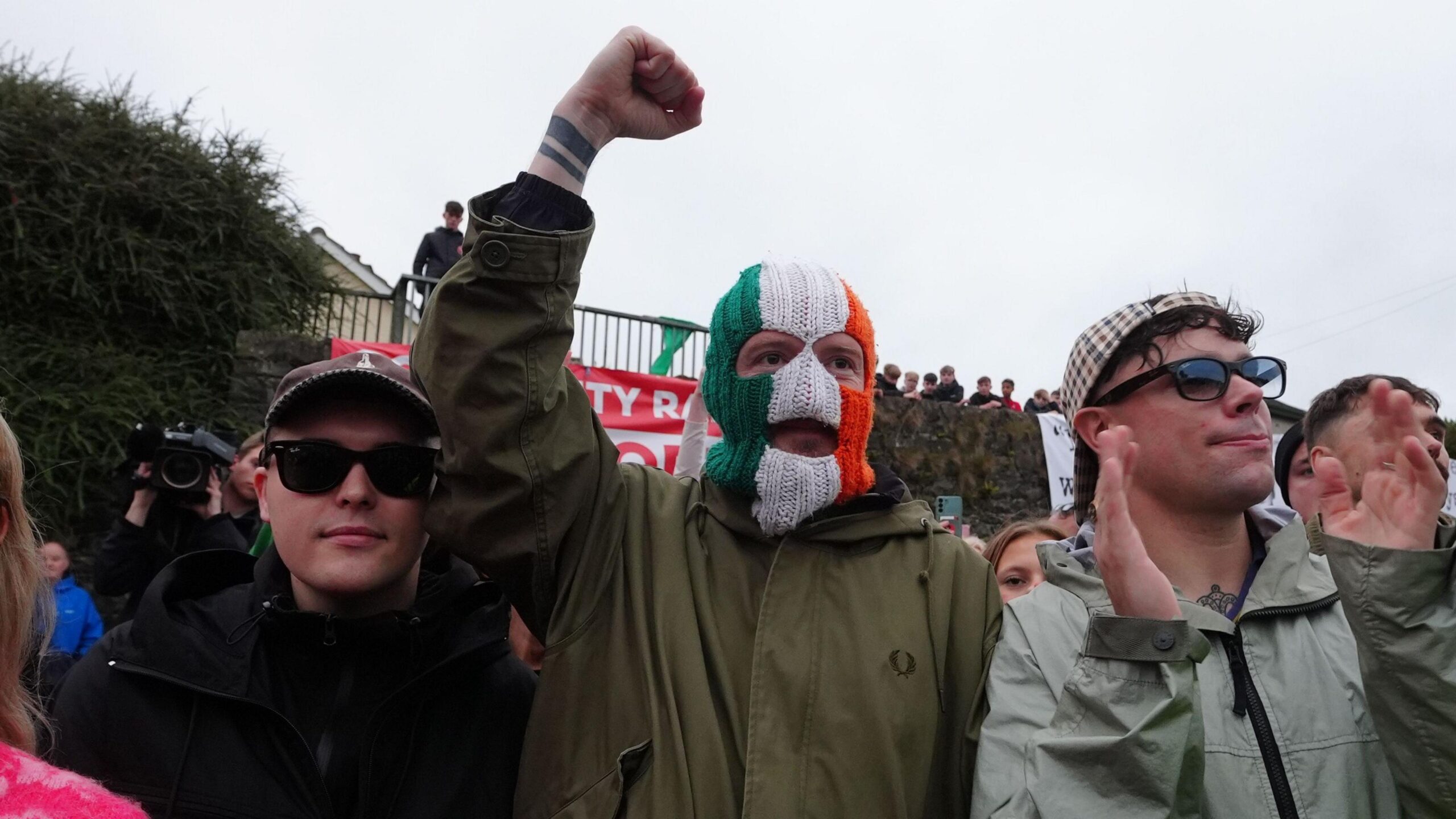 (Left to right) Mo Chara , DJ Próvaí and Móglaí Bap from the band Kneecap stand together in a crowd. Mo Chara and Móglaí are wearing sunglasses and hats. DJ Próvaí is wearing a green, white and orange balaclava with his fist raised in the air.