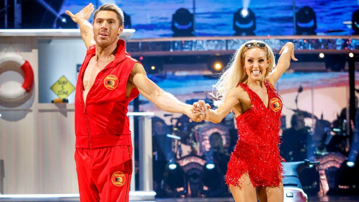 Amy Dowden and Chris Appleton during their appearance on the live show on Saturday 3 October for BBC1's Strictly Come Dancing. Dowden and Appleton wear red Baywatch outfits and are dancing hand in hand on the Stricty dancefloor. Amy has long blonde hair and a red sparkling short dress. Appleton wears a red waistcoat and red joggers and has blonde short hair. They both smile.
