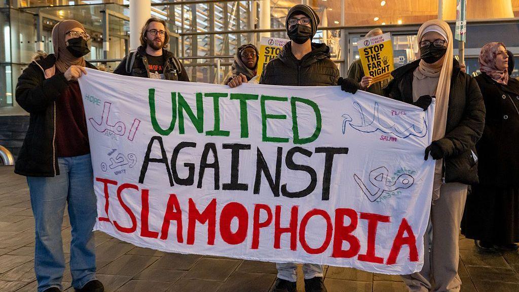 A group of people wearing black face masks with a banner that says "United Against Islamophobia"