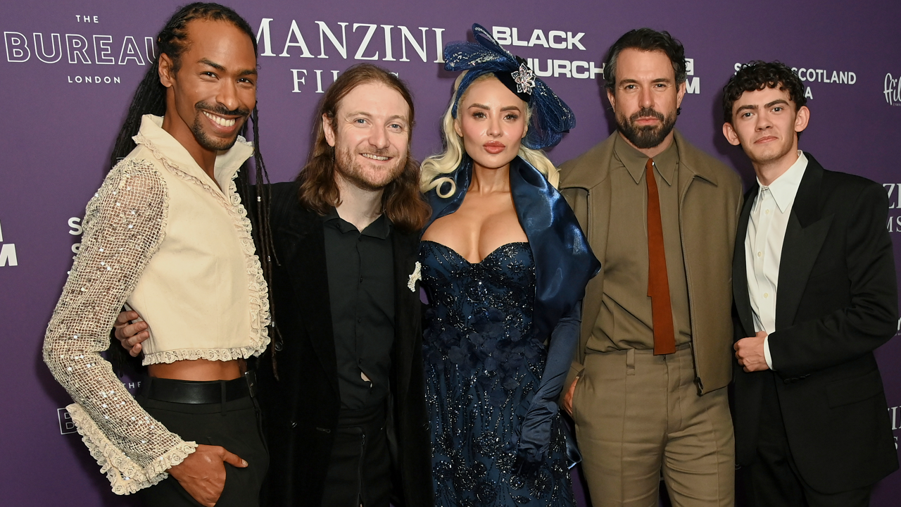 A group of people stood together posing for a photo. On the left a smiling man wears a cream top with sparkly sleeves. The man next to him has a black shirt and black jacket, he has ginger shoulder-length hair. The woman in the middle has blonde hair and pouts, wearing a blue sparkly dress and a blue bow in her hair. Next to her is Tom Cullen who has a brown suit and brown tie and dark black hair. On the right is Joe Locke who wears a white shirt and black suit.