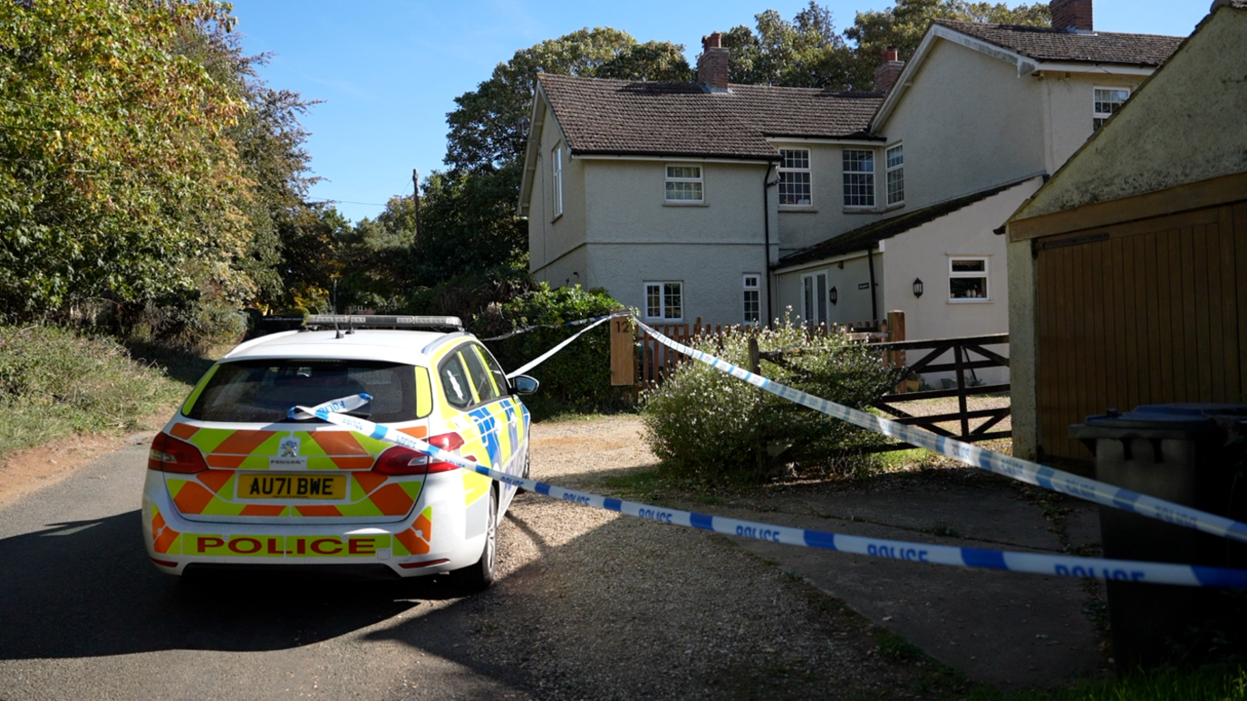 A police car is outside a large house. Blue and white police tape has sealed off the driveway