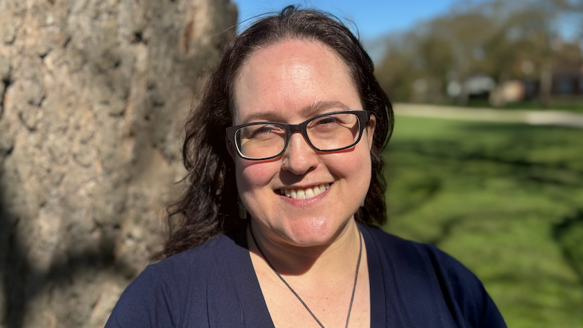 Sam Tipper has brown hair and is wearing a dark blue dress, she also has black glasses. She is standing in front of a tree which is blurred.