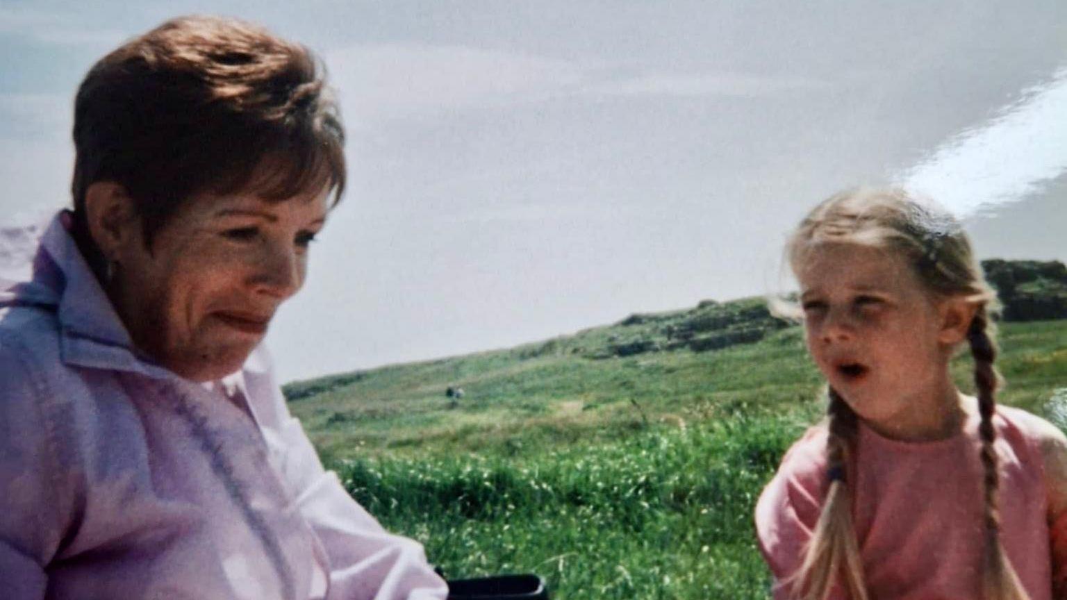 An old picture of Lilly as a young girl with her grandmother. Anne has short brown hair and wears a purple coat, Lilly has long blonde plaits and wears a pink top. There is grass behind them.