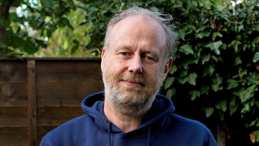 My husband’s death was hard enough – then came the admin Richard Jackson with a beard and short hair wearing a navy blue hoodie with you are enough written on it in white. He has a neutral expression and there is a fence and foilage behind him.