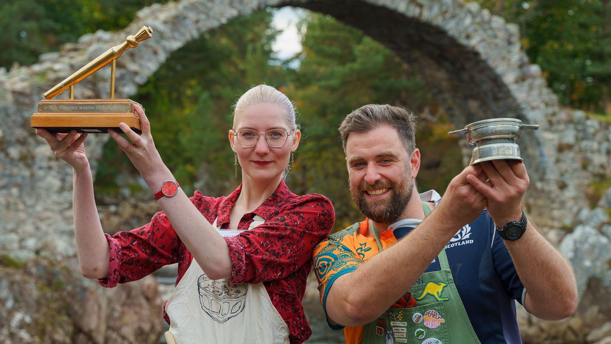 A man and a woman hold up trophies in front of a steeply arched old stone bridge