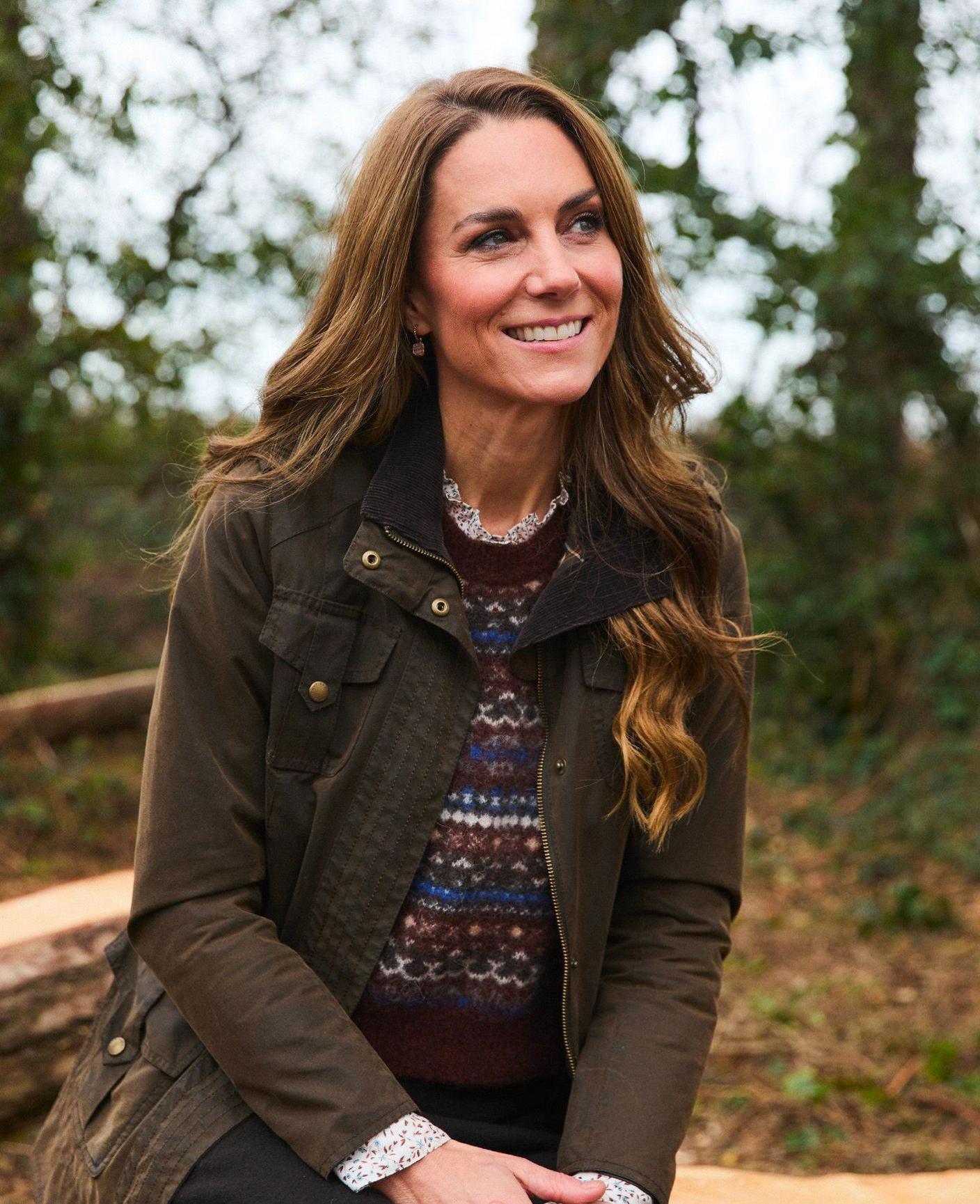 Princess of Wales looks away from camera while posing for a photo wearing a jumper and coat against a forest-like background in an autumn setting.