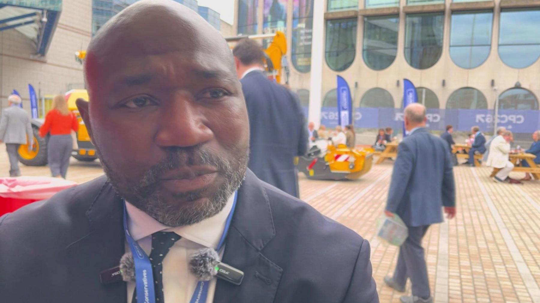 A man in a dark blue suit, white shirt and blue spotted tie with a bald head and a black beard being interviewed.