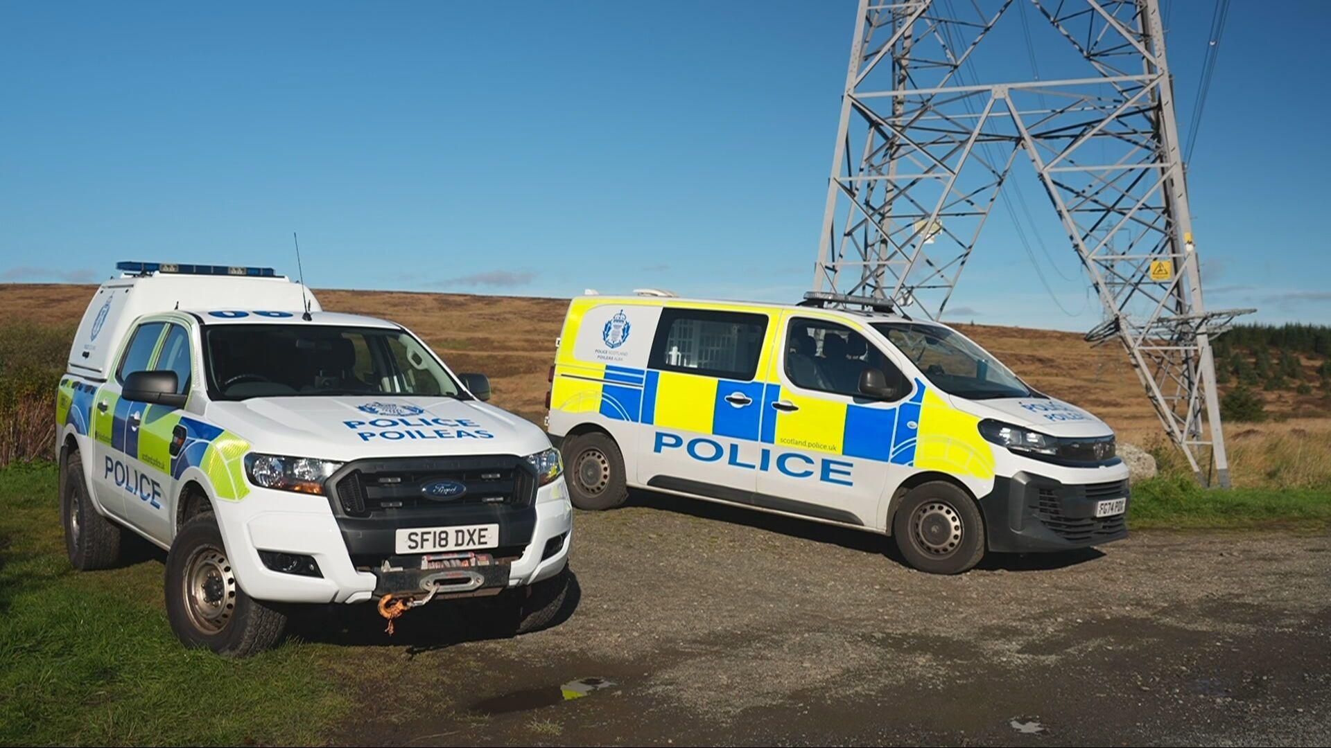 Police vehicles parked in a field by a pylon in daylight
