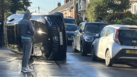 A black Mercedes is on its side in the middle of the road. There is a row of parked cars on the right hand side and a person in a grey sports tracksuit is standing in front of the car and is speaking into his phone.