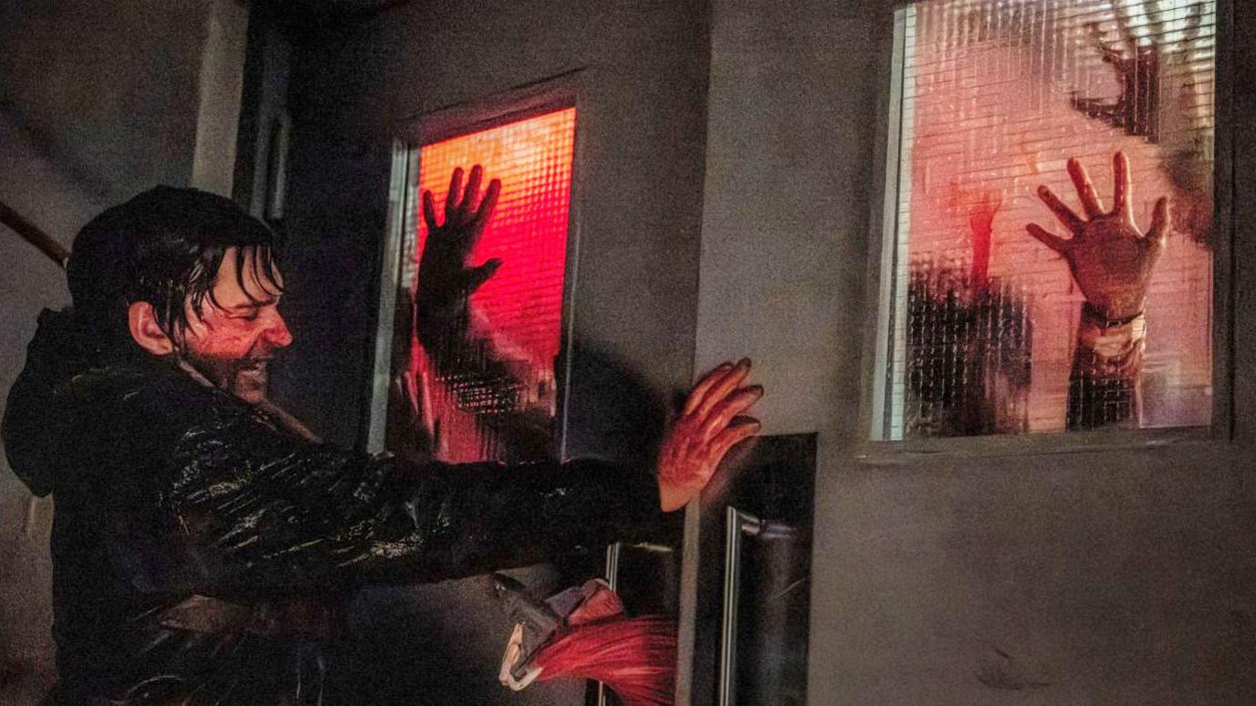 Still from Resident Evil shows Bryan, played by Austin Abrams, desperately pushing against a set of double doors in a hospital hallway. His hands and face are covered in blood, which also matts his hair to his head. A small crowd of zombies can be seen through frosted panes in the doors, their hands pressed against the glass.