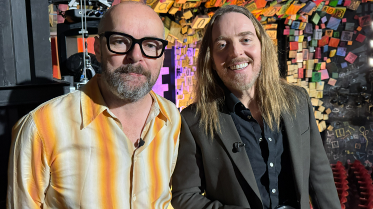 Dennis and Tim smile at the camera in the stage area in front of a multi-coloured background. Dennis has a yellow shirt on with a beard and glasses and Tim is in a dark coloured shirt and jacket with long hair and a beard.