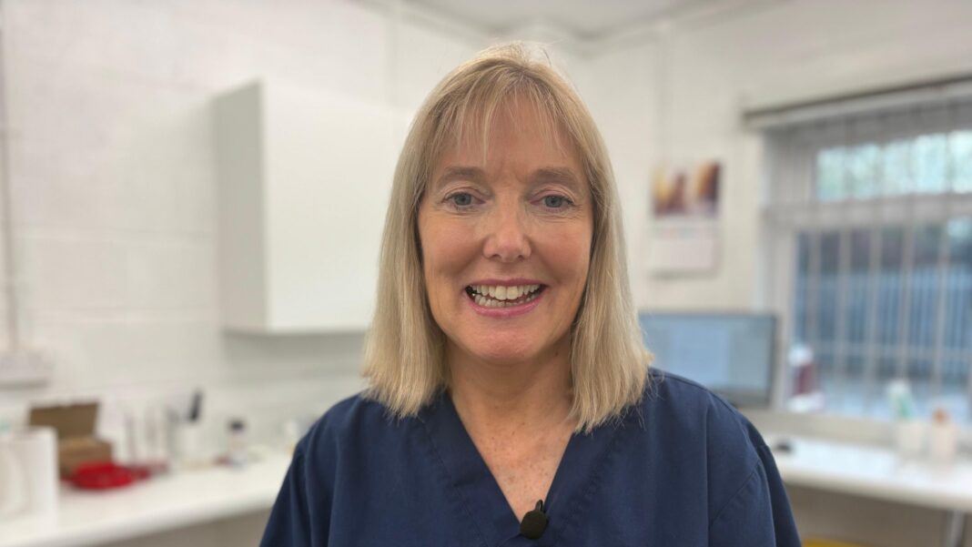 Chronic UTIs made MP ‘feel like a drug user’ A female doctor with blonde shoulder length hair smiling at the camera. She is in a laboratory where urine is tested for signs of infection. She is wearing a blue top.