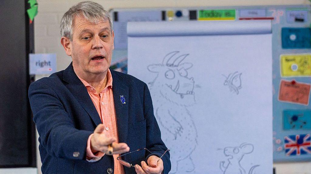 A man with grey hair, peach shirt and blue jacket, holding a pair of glasses, stands in front of a white drawing board.