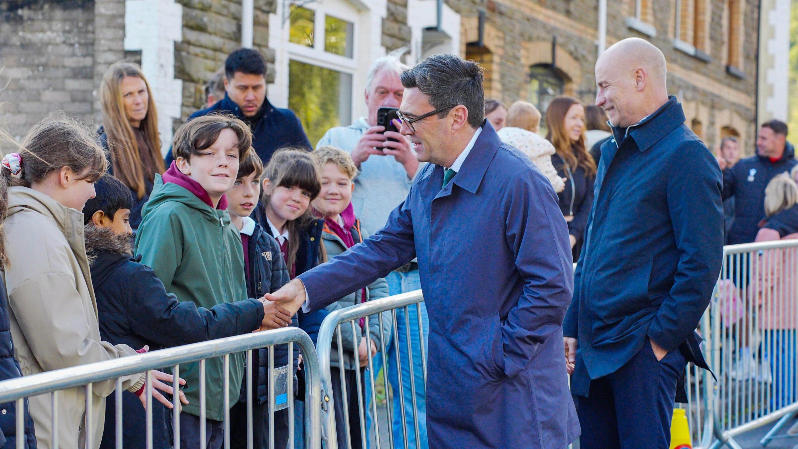 Andy Burnham in a long navy coat shaking hands with children next to a grey metal barrier on the side of the road.