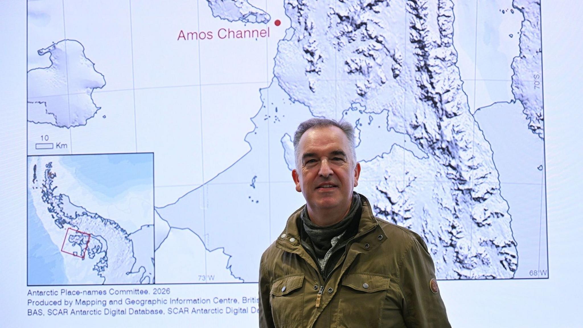 A man is in the centre of this image. He is smiling and looking directly at the camera. He's wearing a khaki jacket over a dark fleece and scarf. The background is a map of the Antarctic Peninsula with the Amos channel marked with a red dot and label.
