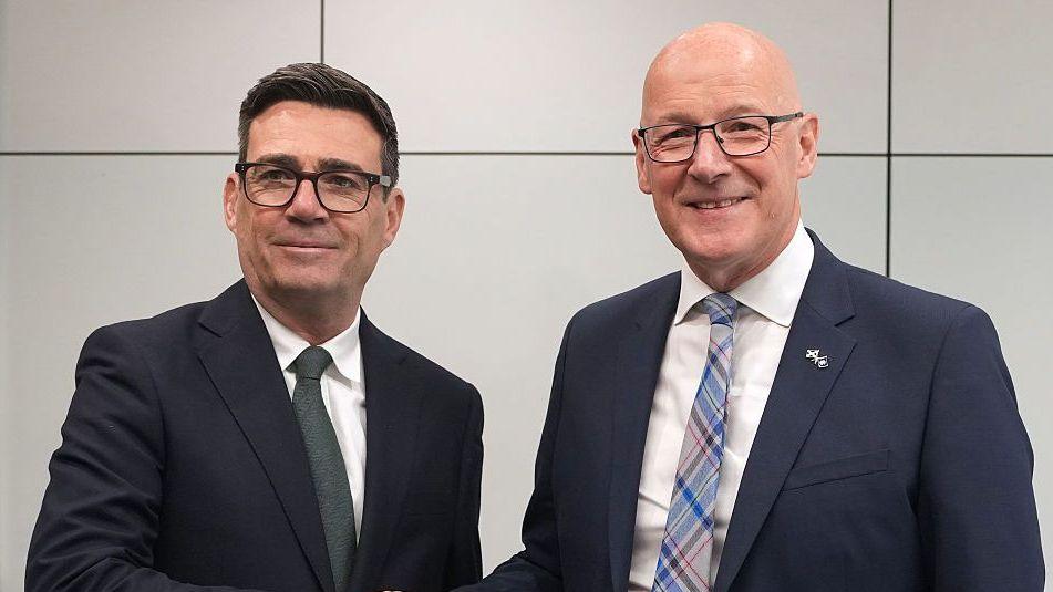 Two men, one with dark hair and one bald, smile as they stand alongside each other. They are each wearing a dark jacket and white shirt with the bald man wearing a tartan tie and the other man wearing a green tie