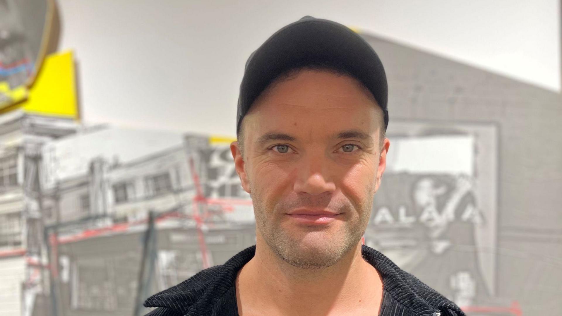 Artist Endless wears a dark baseball cap and black shirt, and stands against the black and white backdrop of his exhibition.