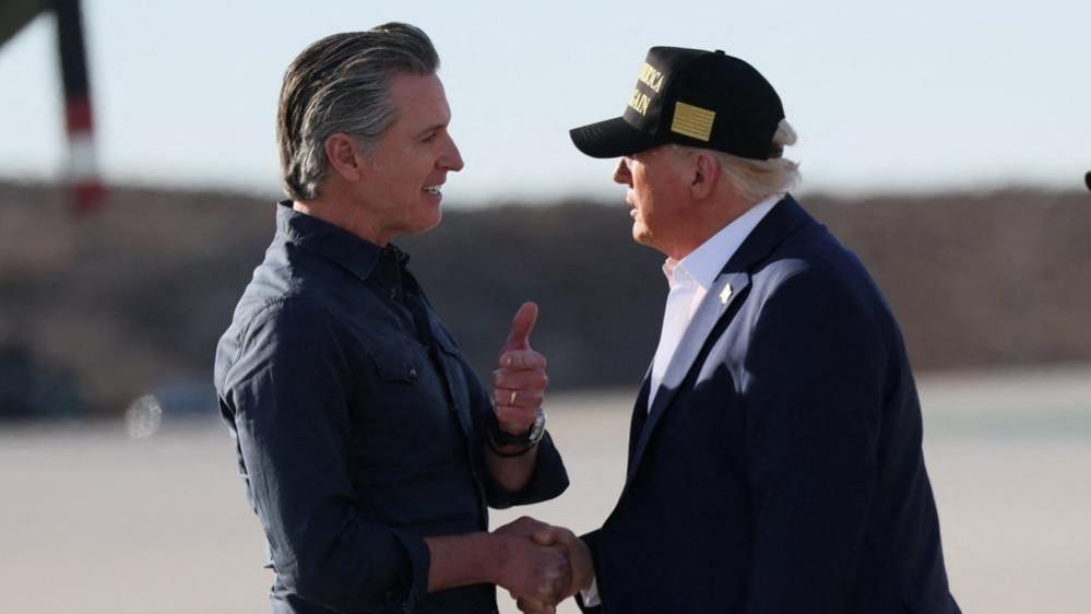 California Governor Gavin Newsom and US President Donald Trump shake hands on airport tarmac. Trump, on the right, wears a baseball cap.