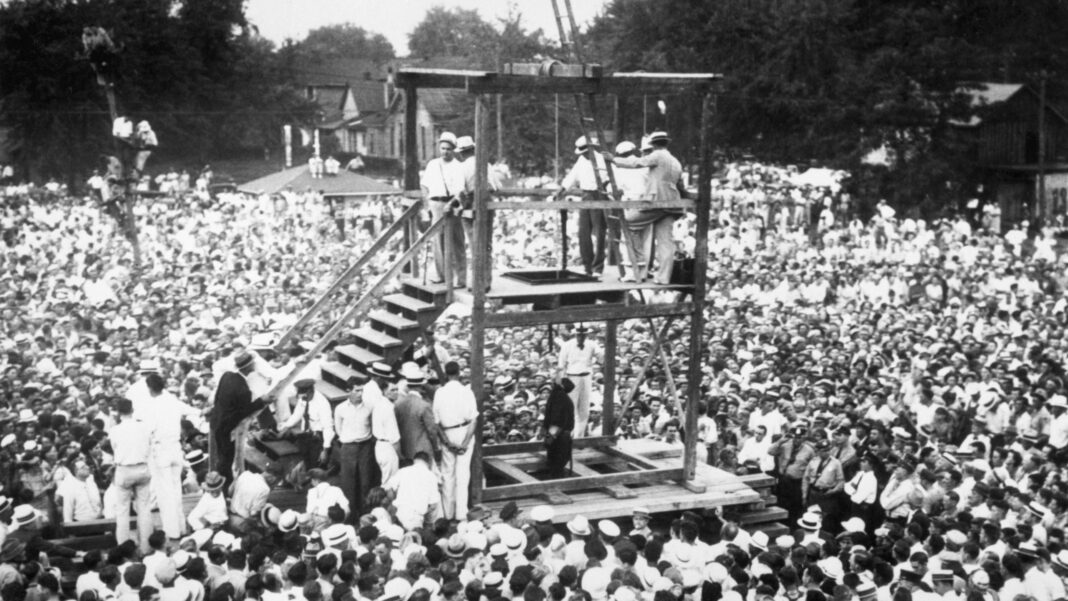 Men stand on wooden gallows, looking through a trap door at a black-clad figure hanging from a rope, as throngs of onlookers surround them
