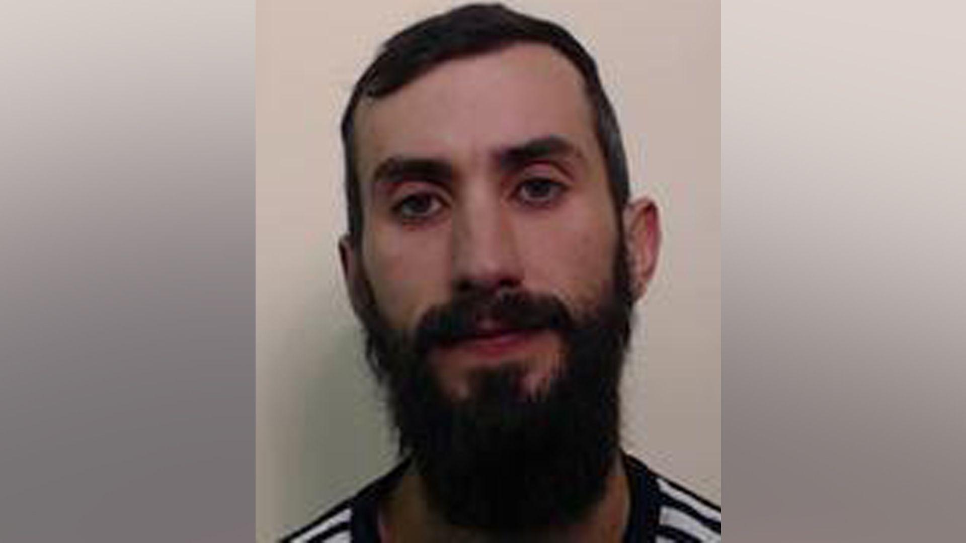 A mugshot of a dark-haired man with a thick beard. He is looking directly at the camera.