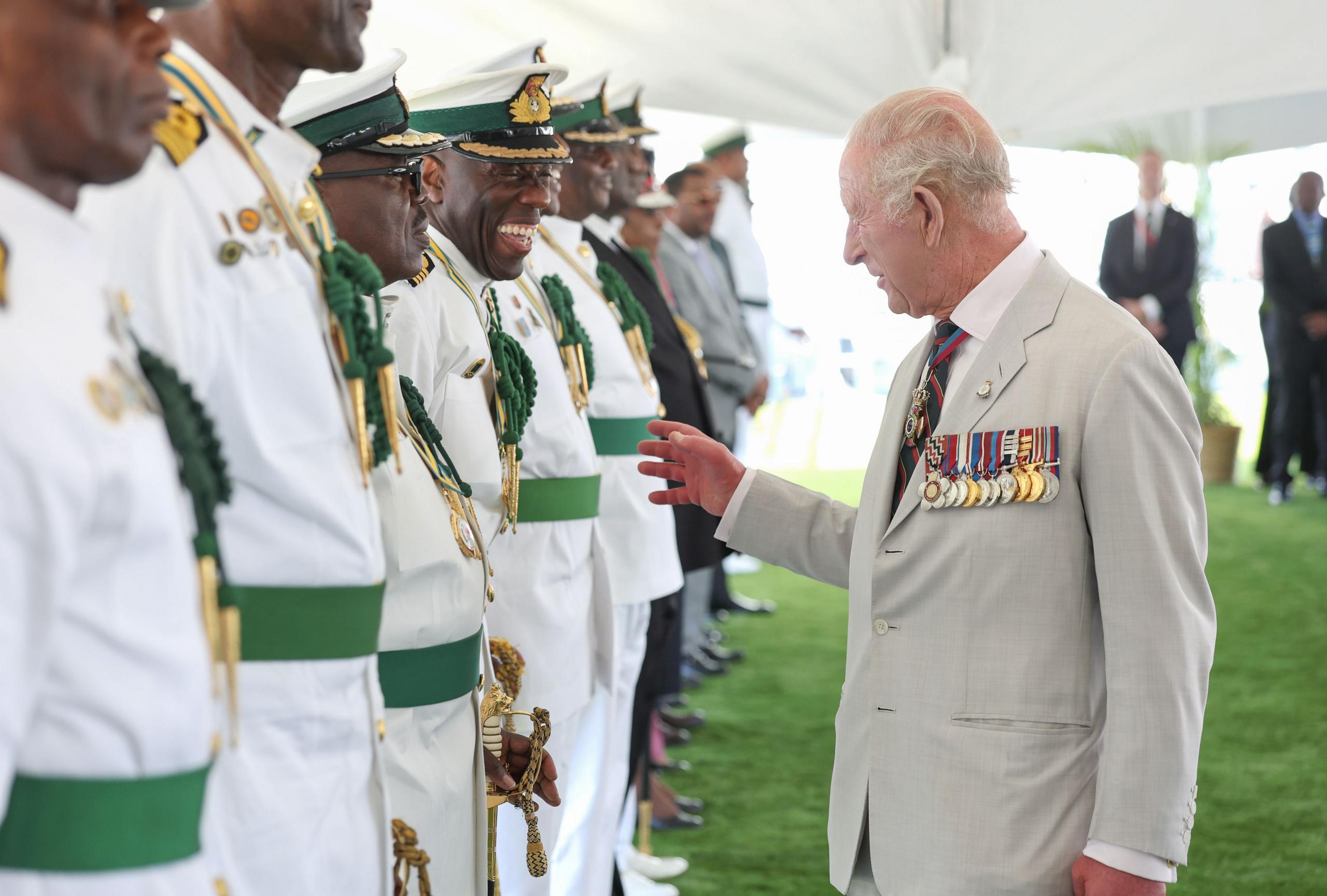 King Charles, wearing a light beige suit with military medals displayed speaks to veterans wearing white uniforms highlighted with green