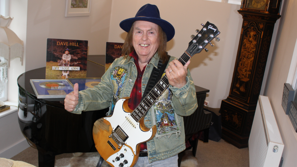 A man in a black hat and green patterned jacket is holding a guitar and smiling at the camera with his thumb up. He is standing in a room with a piano and records and framed discs on top.