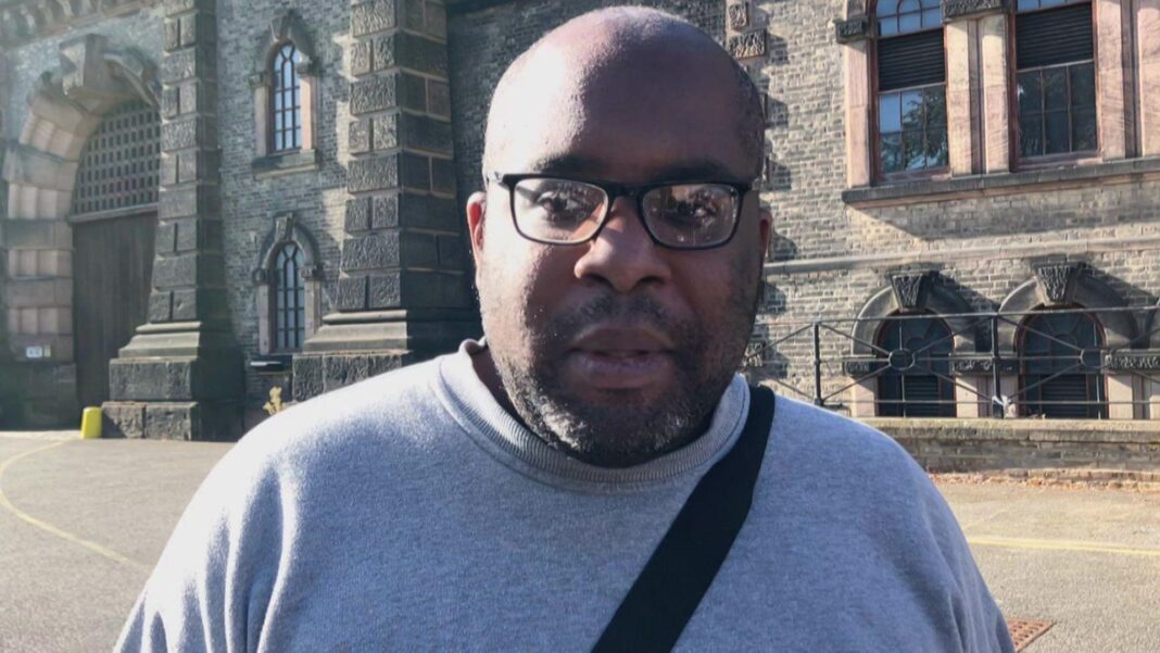 Close-up of Michael Bogle wearing black-rimmed glasses, a grey crewneck jumper, and a black cross-body bag strap, standing outside the stone entrance of a prison building.