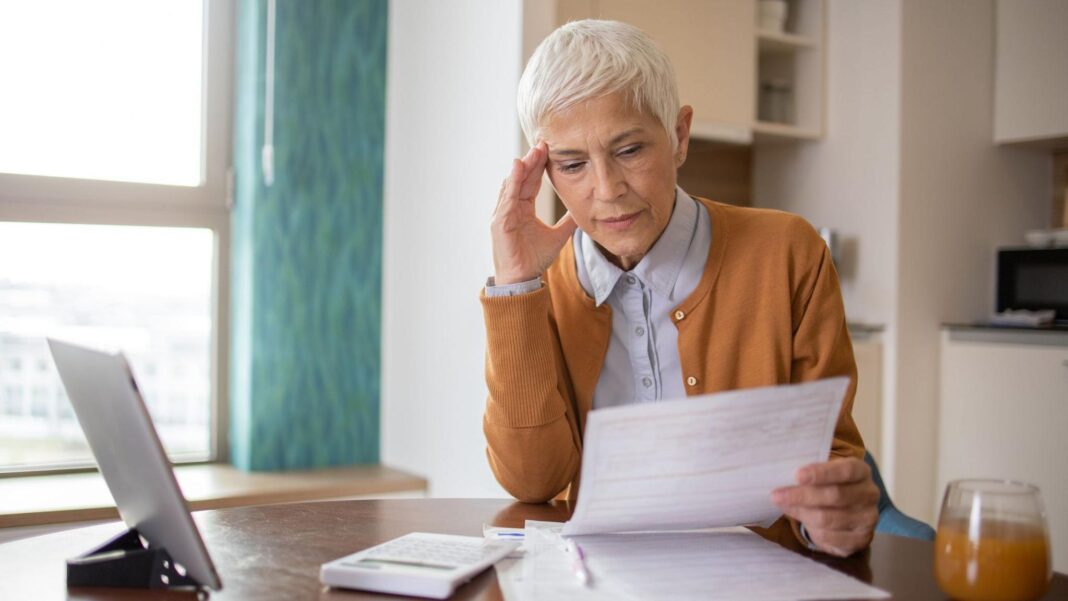 A woman with grey hair sits at a table looking at a bill. On the table there is a calculator and tablet.
