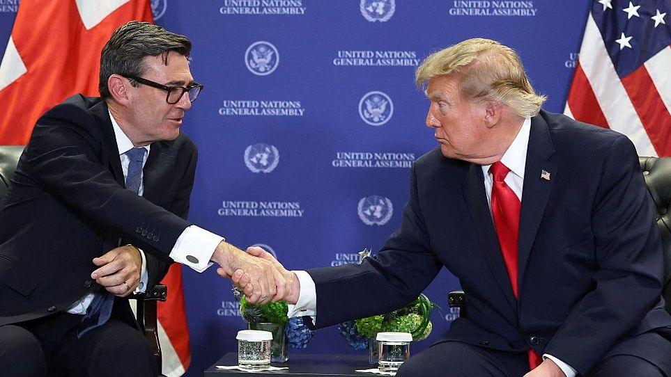 Andy Burnham and Donald Trump sit on chairs on either side of a small table. They are leaning towards one another and shaking hands. Both are wearing dark suits: Burnham is wearing a blue tie and Trump is wearing a red one. A background with the words 'United Nations General Assembly' can be seen behind them.