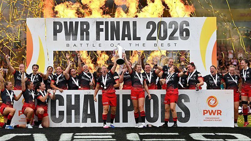 Saracens players, wearing black tops and red shorts, lift the PWR trophy