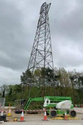 A tall electricity pylon, surrounded by building site equipment, tape and cones. Trees are visible behind it.