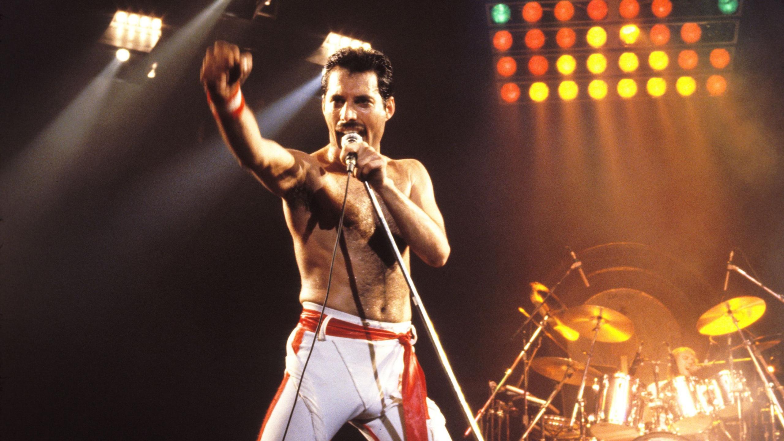 Freddie Mercury is pictured on a stage singing, with a microphone to his mouth and his arm raised. He is wearing white trousers. Behind him is a drum kit and lights.