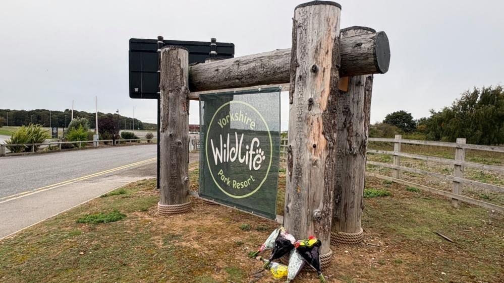 The park sign with some flowers laid by it. The sign is green fabric hung on a large wooden frame. There is grass all around. There are bunches of flowers at the scene.