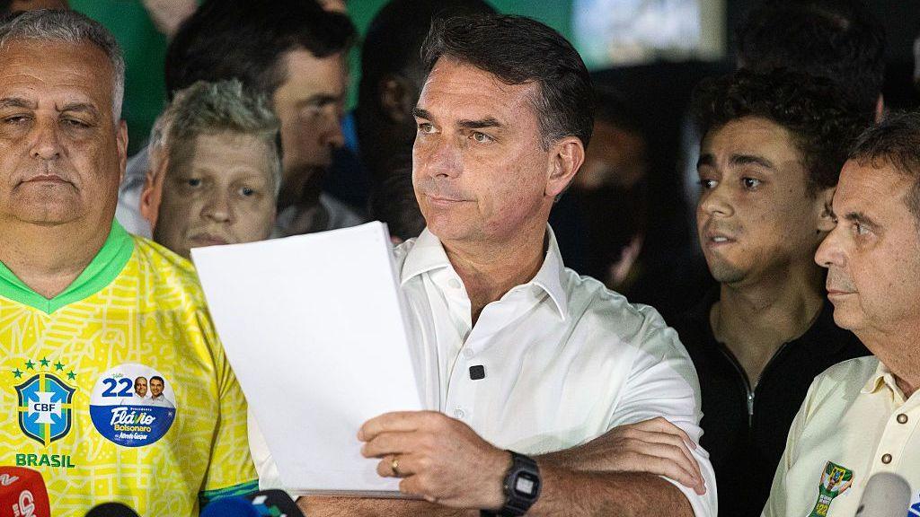 Bolsonaro standing while holding a wad of paper. He is surrounded by people who do not look happy