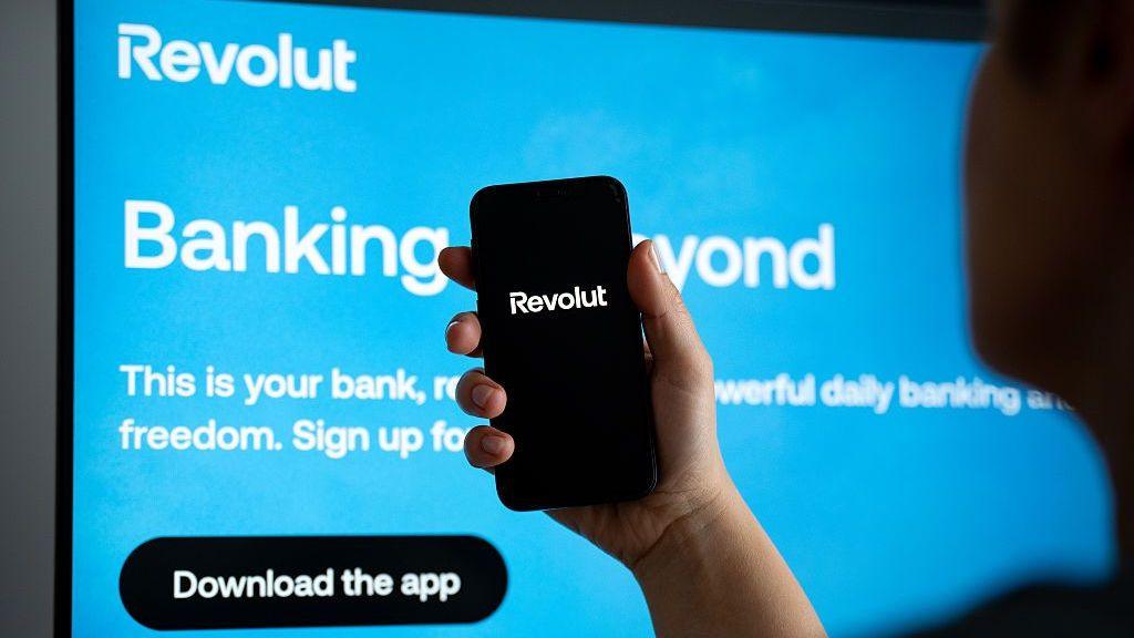A person holding a smartphone displaying the Revolut app logo, in front of a display showing a bright blue Revolut advertisement.