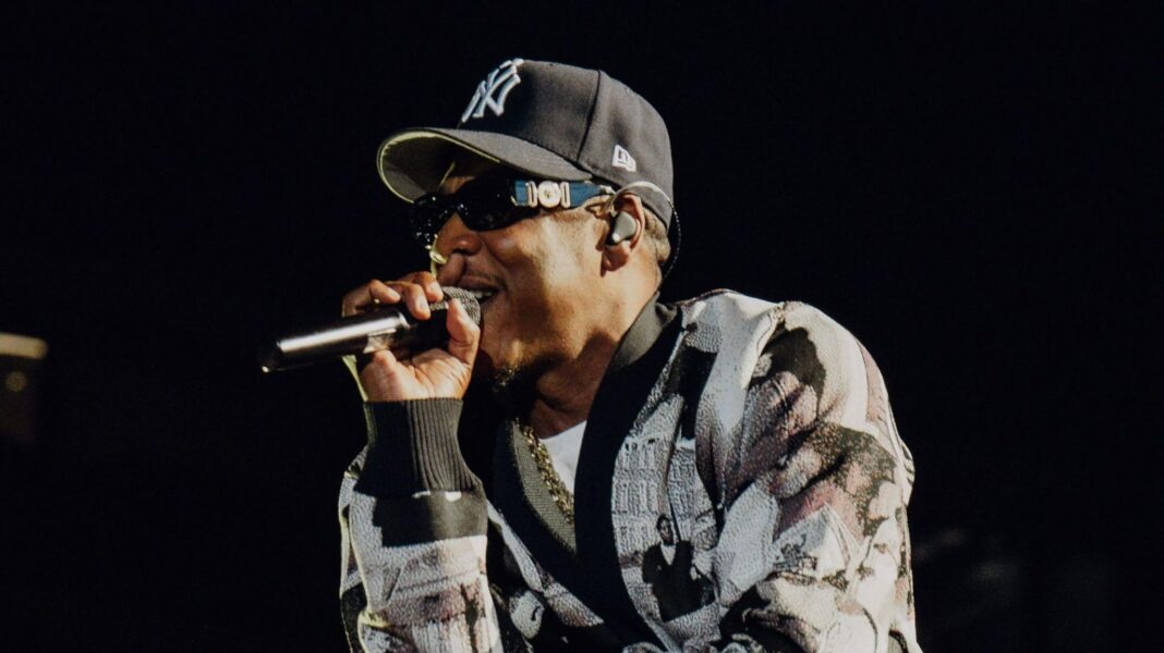 Jay-Z raps into a microphone while performing on stage. He wears a black and white cardigan and a New York Yankees hat.
