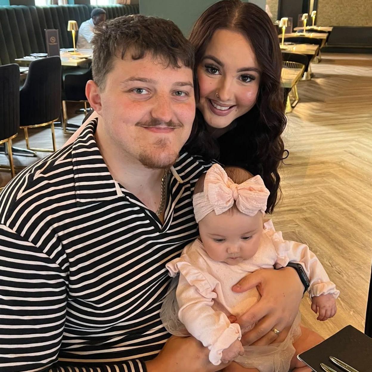 Sophie and Ryli Price are sitting in a restaurant with their baby daughter Florence. Ryli has a black and white horizontal striped shirt on and has short light brown hair with a goatee. Sophie is to his left and has long, dark wavy hair and a nose piercing. Florence is on her dad's lap and is wearing a light pink jumper with frilly cuffs and a matching bow on her head