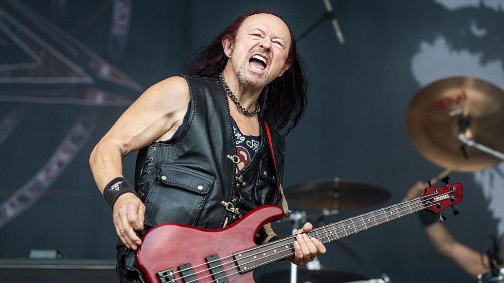 Conrad Cronos Lant on stage with Venom at the Sweden Rock Festival in June 2026. He is bald at the front but has really long, dark reddish brown hair flowing down from the top of his head. He has his mouth open as if shouting but his face suggests happiness. He also has a short, stubbly goatee beard and moustache with flecks of grey. He is wearing a black leather waistcoat with a really big buttoned pocket and laces tied at the side seam. Under that is a black vest which seems to be a Rolling Stones T-shirt that has had the collar and sleeves cut off. He also wears black leather trousers, a thick bronze coloured link chain round his neck and a black sweatband on his wrist. He is playing a blood red and black coloured bass guitar and behind him are symbols on one side of a drum kit with the drummer's right arm in the air.