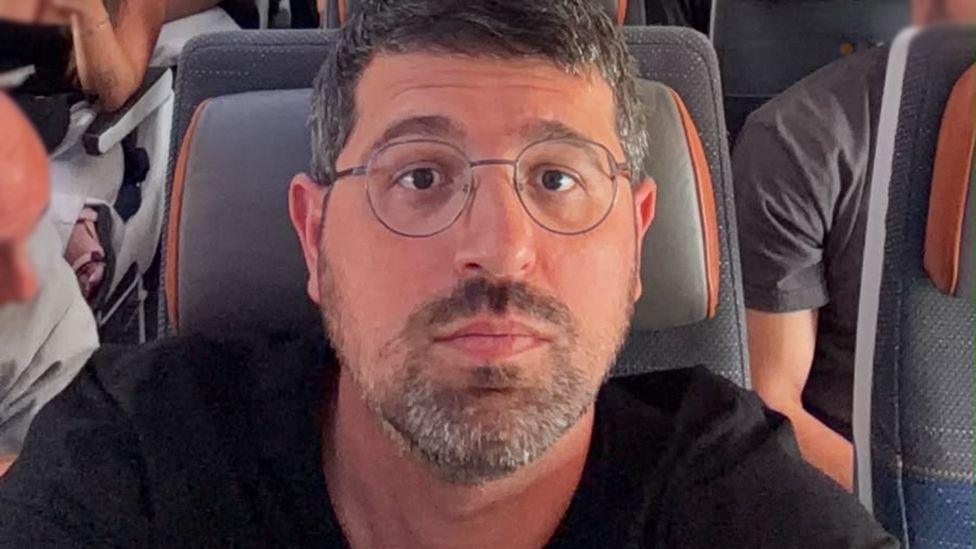 A selfie of Adam Polovincik sitting on a busy aeroplane with a grey and orange seat cushion. He has short dark hair and a beard and is wearing glasses and a black t-shirt