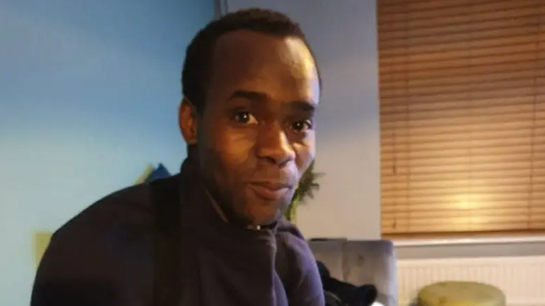 Anton Green in a dark high-neck top sits indoors, facing the camera. The room has soft lighting, with wooden blinds covering a window on the right and a chair and cushions visible in the background.