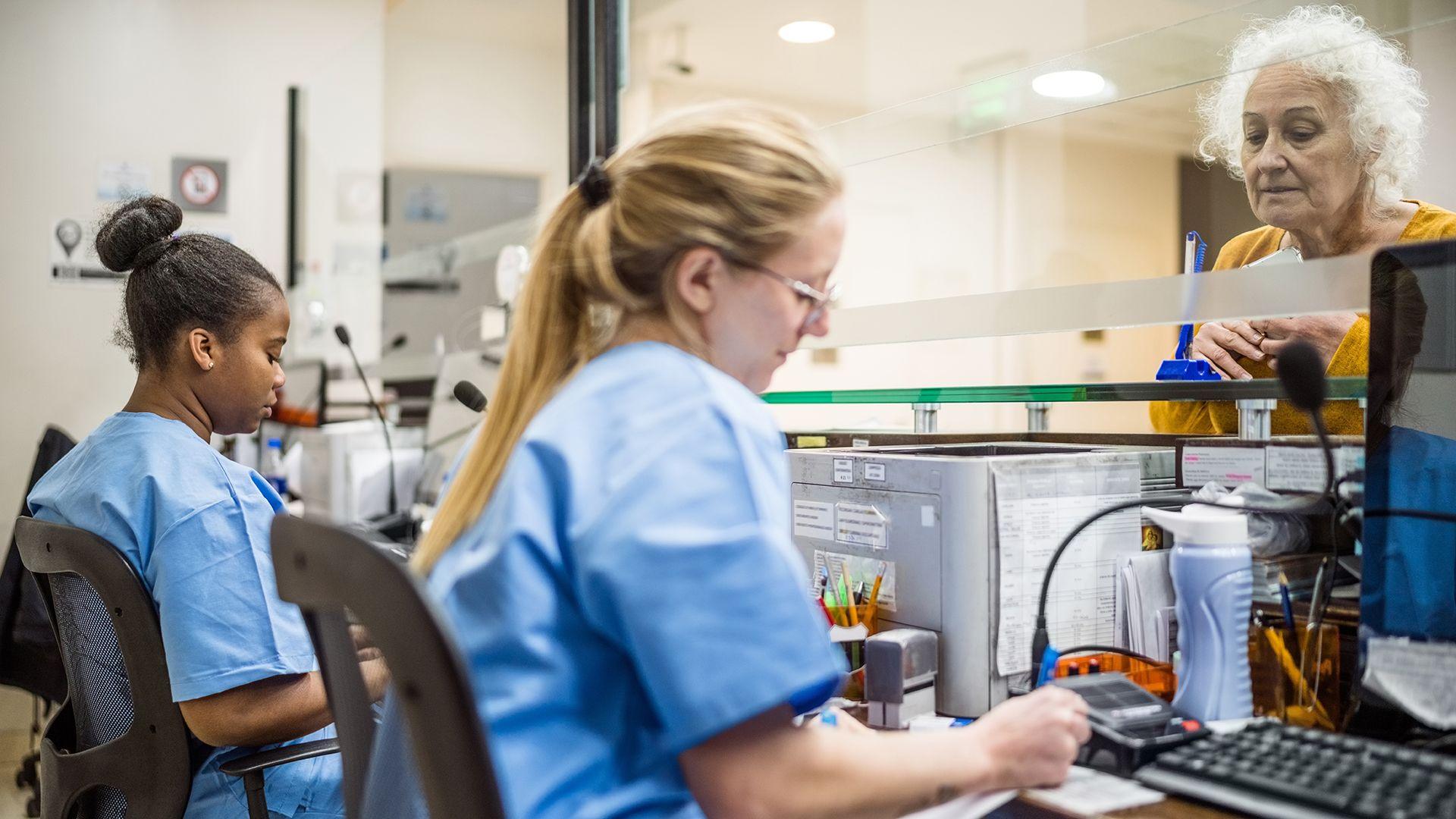 Stock photo shows two NHS receptionists sitting at a desk in front of computers behind a glass pane with a hospital visitor on the other side of the pane being attended to by one of the staff, in a hospital setting.