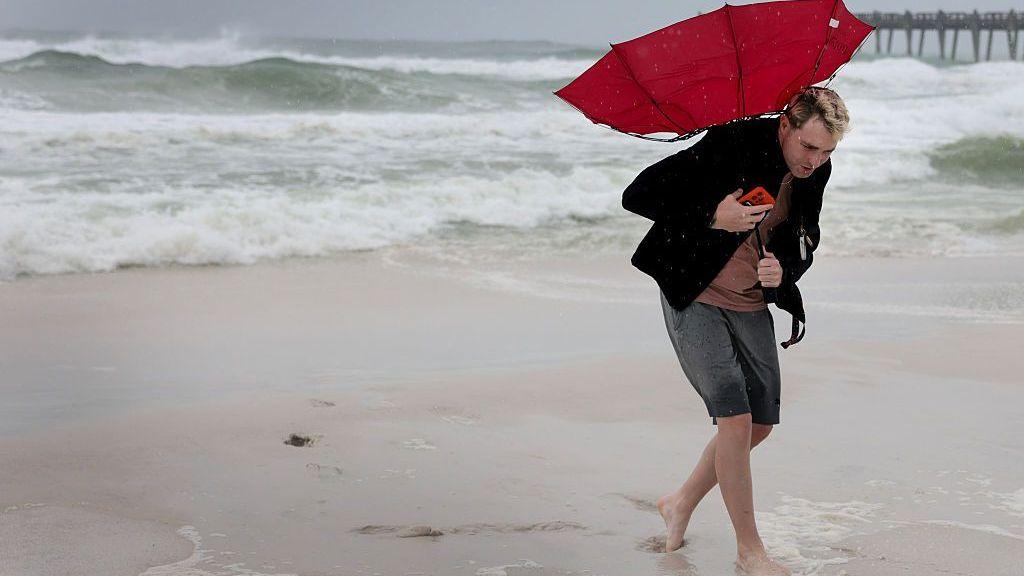 Michael Collins walks on a beach with his red umbrella turned inside out as winds pick up ahead of Hurricane Isaias on 9 October 2026, in Pensacola Beach, Florida.